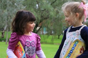 Two little girls of different background smile at each other holding books.