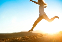 Women leaping across a field with sunshine behind her. 