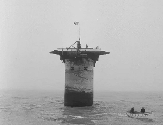 Black and white photo of concrete pillar looming out of the sea, with row boat in the foreground