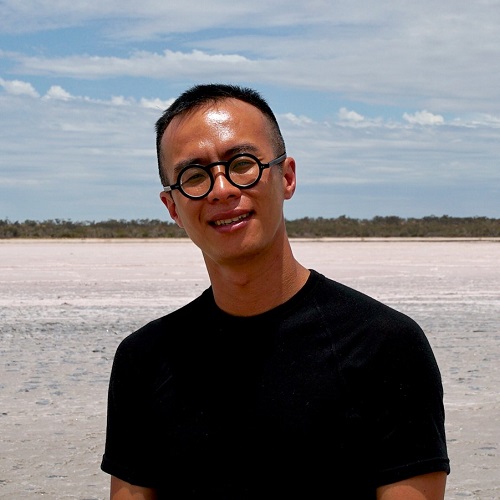 Portrait of James Nguyen, a Vietnamese male wearing black, round-framed glasses and a black t-shirt, standing in front of a salt lake.jpg