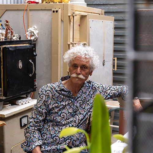 Bruce Nuske, a Caucasian male with white curly hair and moustache, sits in his studio wearing a blue paisley print shirt.jpg