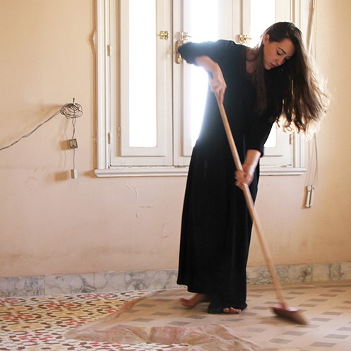 Still image from Dana Awartani's moving image work. The artist, an Arabic woman with long dark hair wearing a long black dress, stands in an abandoned house sweeping away coloured sand that has been laid on the floor to create the effect of tessellated tiles.jpg