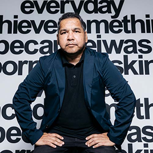 Photo of Vernon Ah Kee sitting in front of one of his text based works. Vernon is an Aboriginal man with short dark hair wearing a navy suit jacket over a black shirt and dark pants.jpg