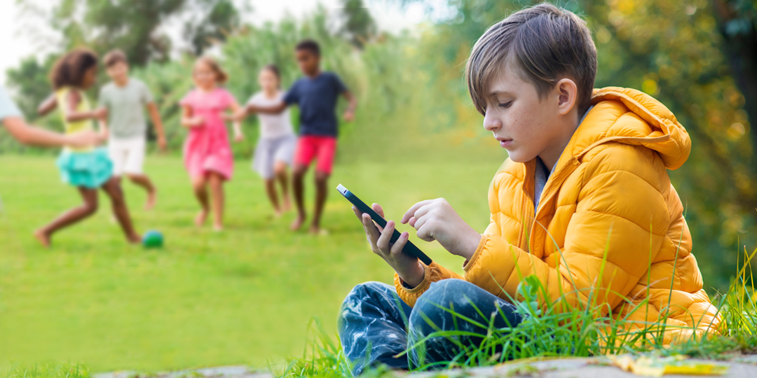 Young boy playing on phone while his friends play soccer